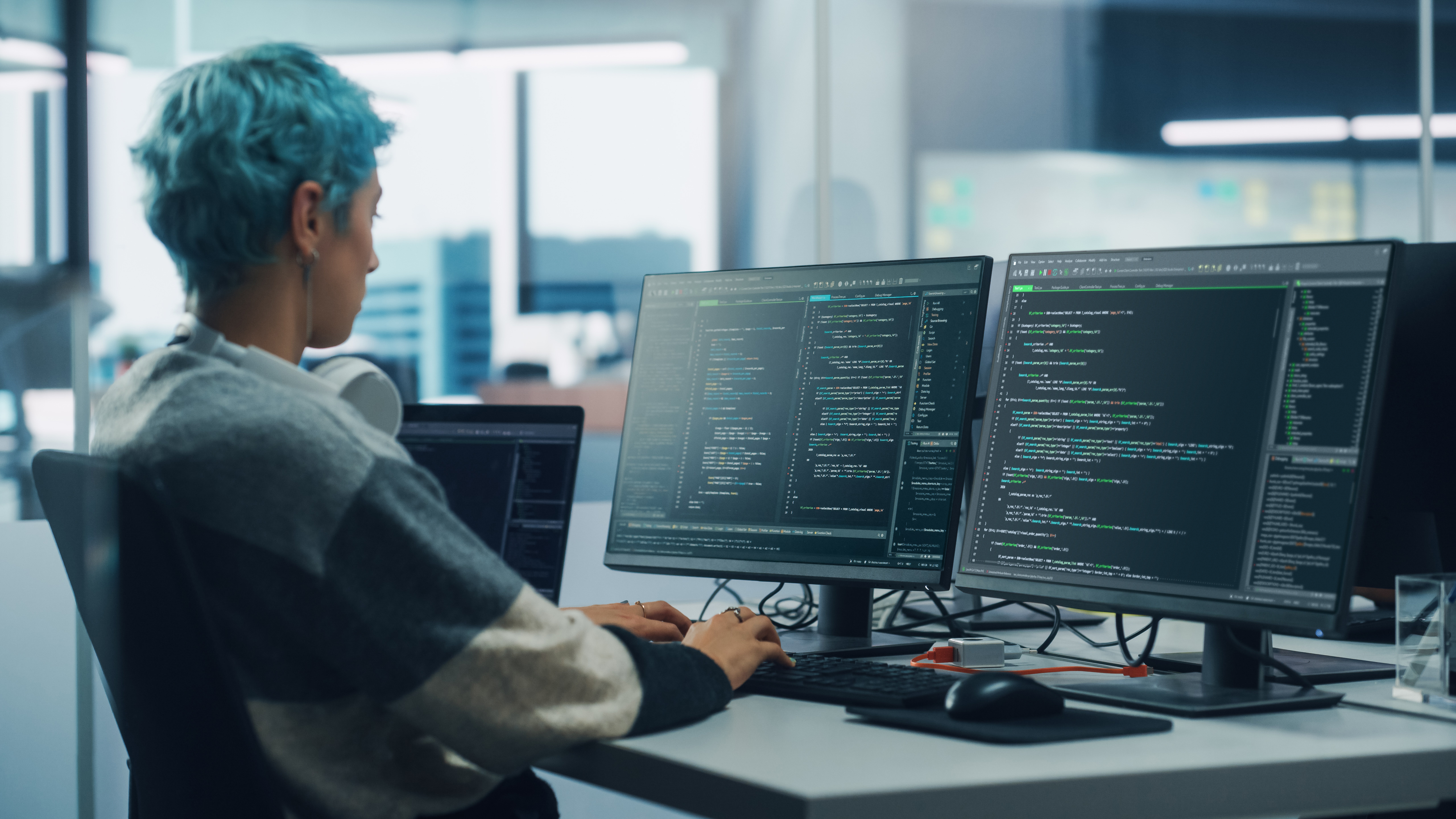 Programmer with blue hair sits at their desk coding across two screens